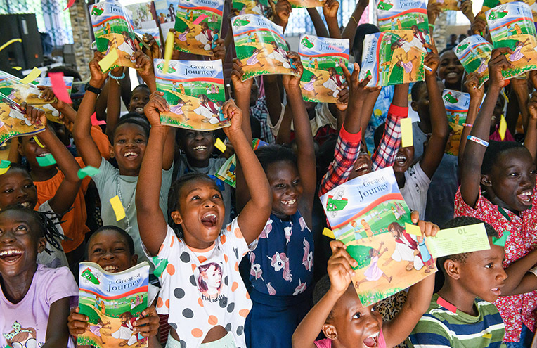Children joyfully holding up copies of “The Greatest Journey” booklets during a celebration event.