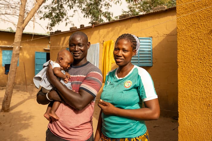 Pastor Ernest stands proudly with his wife and their baby outside their home, a picture of a healthy and hopeful family transformed by the gift of sustainable agriculture.